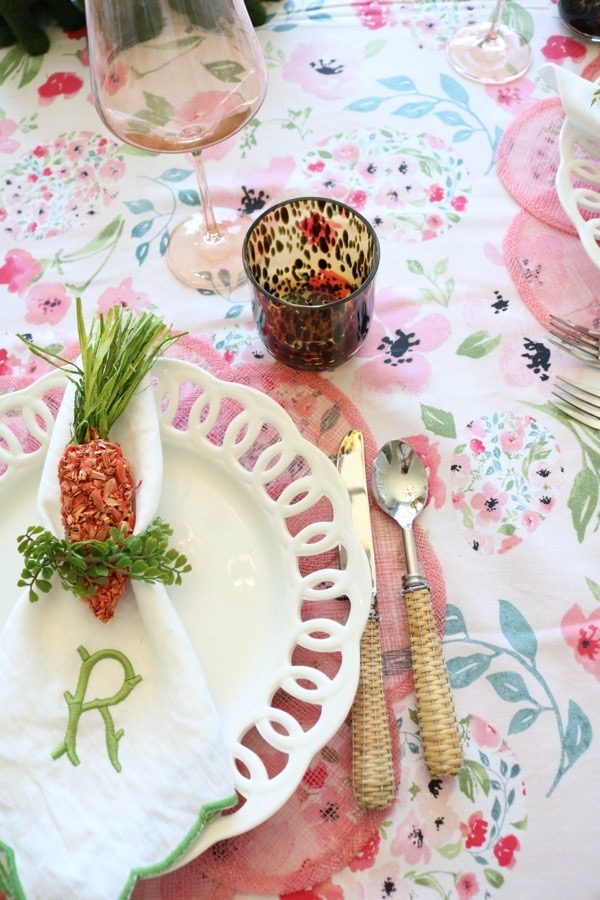 A floral Easter egg tablecloth is layered with pink placemats and white dishes with a carrot tucked into the napkin.